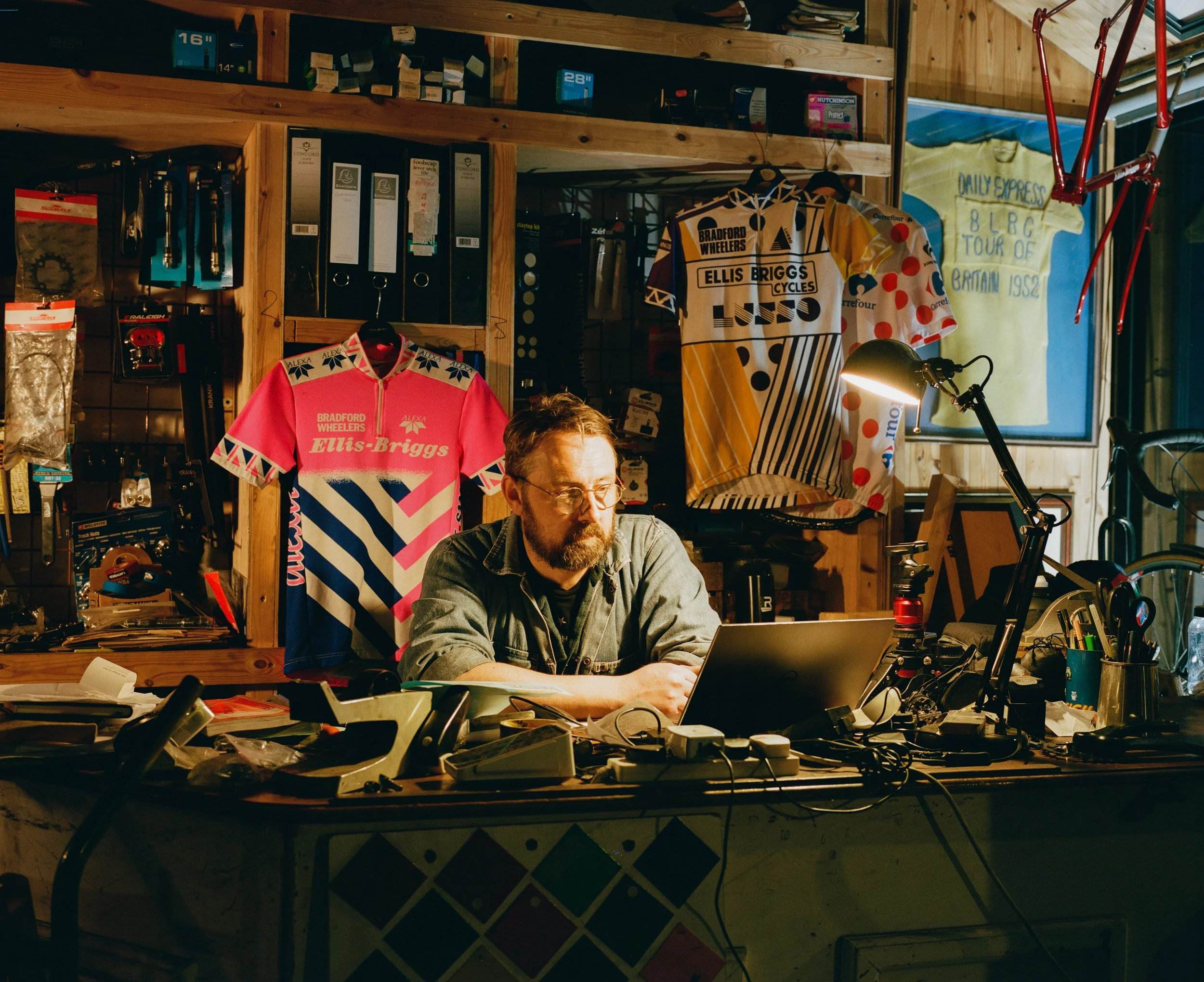 A man with glasses and a beard working on a laptop at a cluttered desk inside a bicycle shop, with cycling jerseys hanging on the wall behind him and bike parts and equipment scattered around.