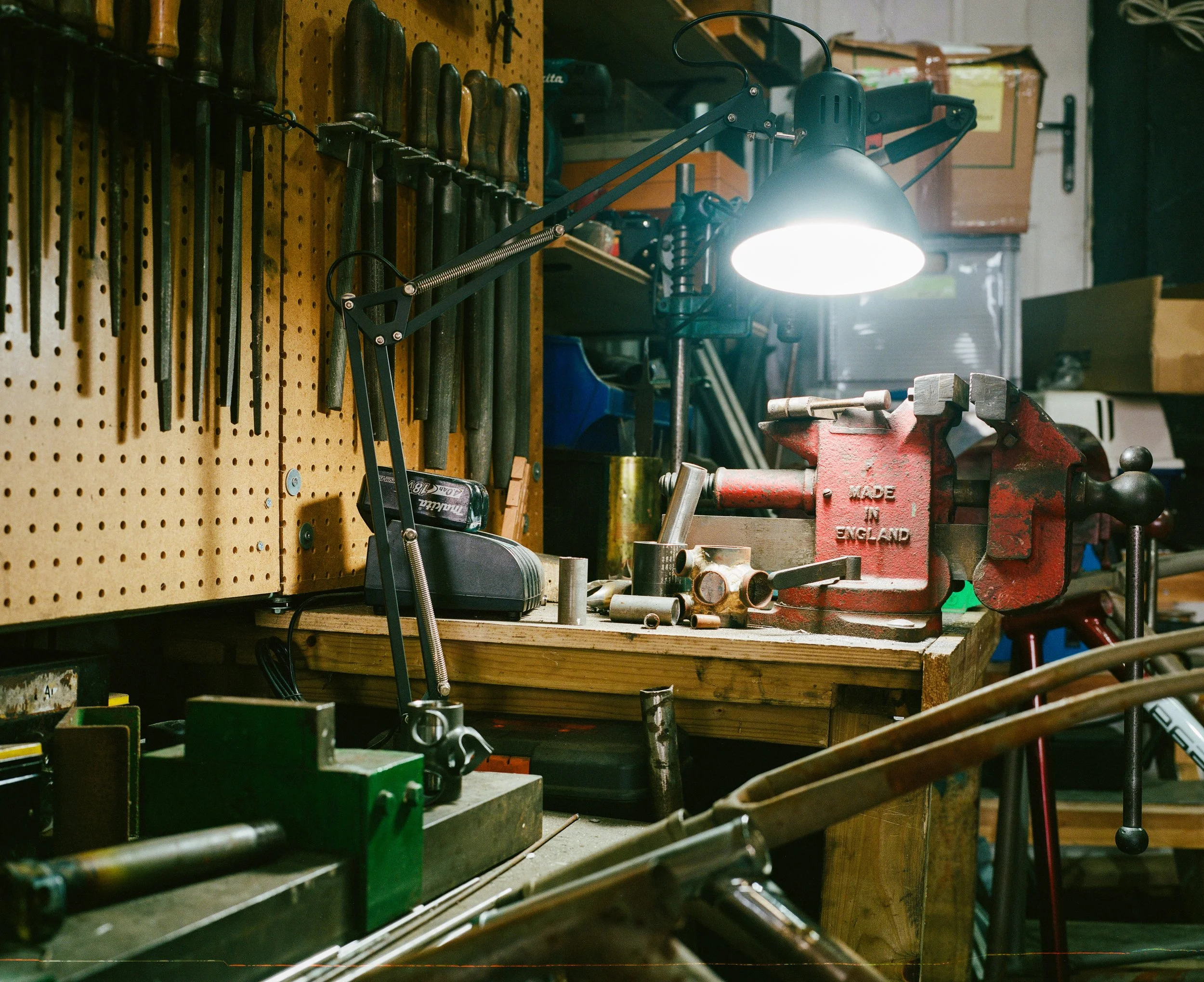 A workshop workbench with various tools, a red vice, and a lamp illuminating the area. Hand tools are hanging on a pegboard on the wall.