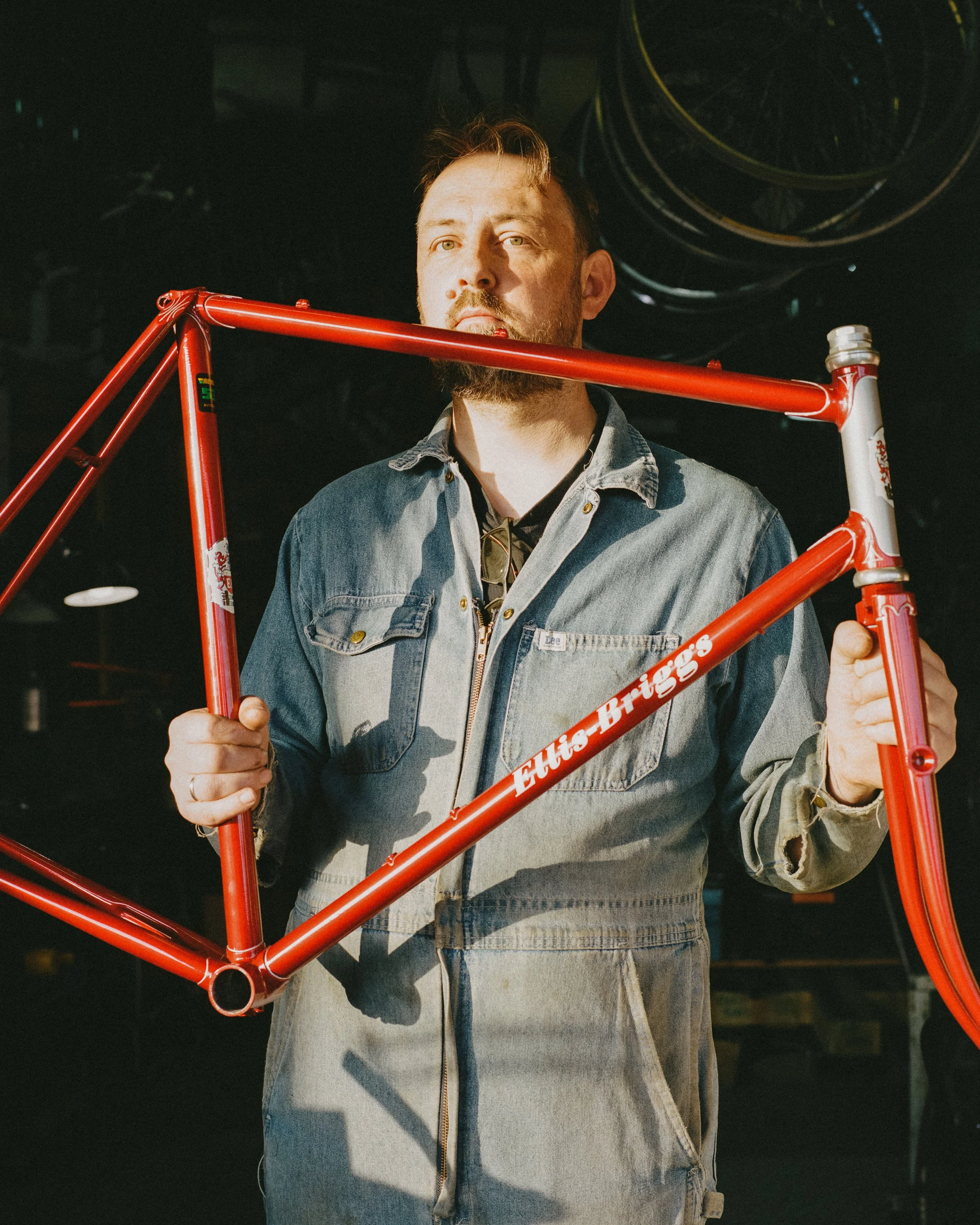 A man with a beard and wearing a denim jacket is holding a red bicycle frame, standing in a workshop or studio with dark background and industrial lighting.
