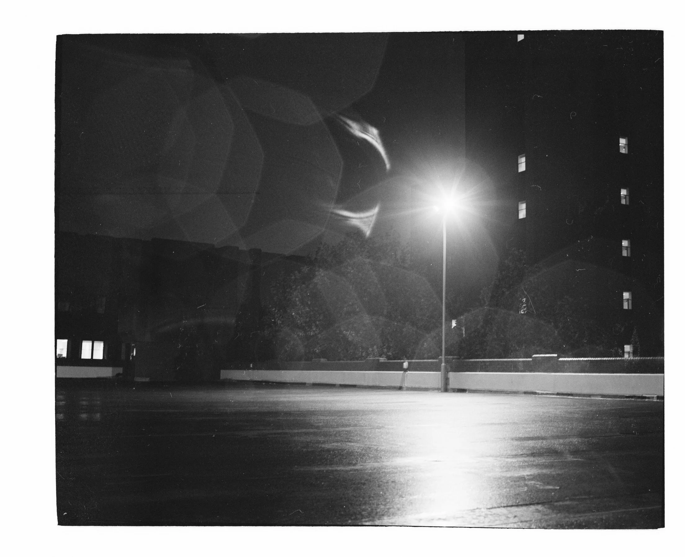 Black and white photograph of a building at night with a bright light, possibly the moon, in the sky and a streetlamp illuminating the ground below.