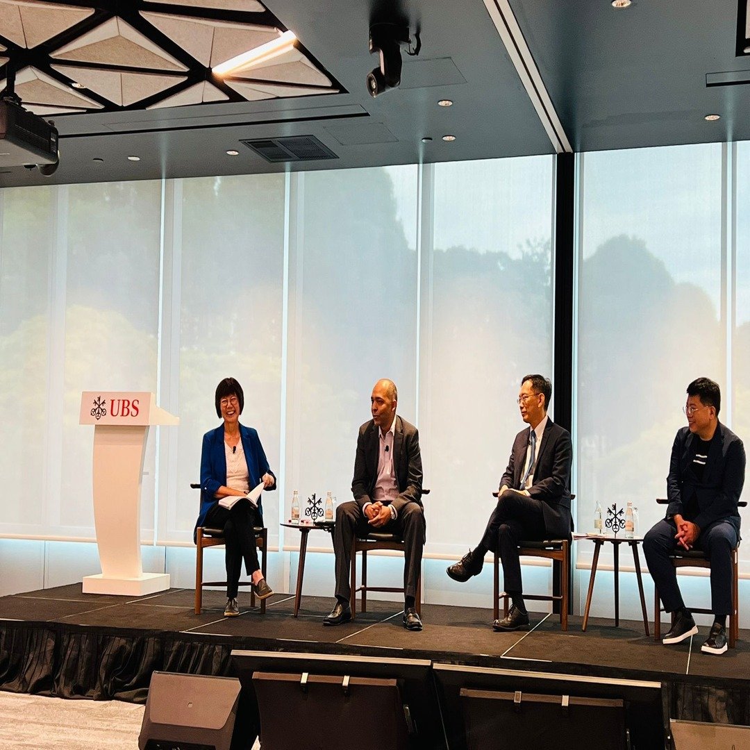 Four people sitting on a stage at a UBS event, engaged in a panel discussion, with each person seated in a chair. There is a podium with the UBS logo on the left. The background features large windows with a view of blurred greenery.