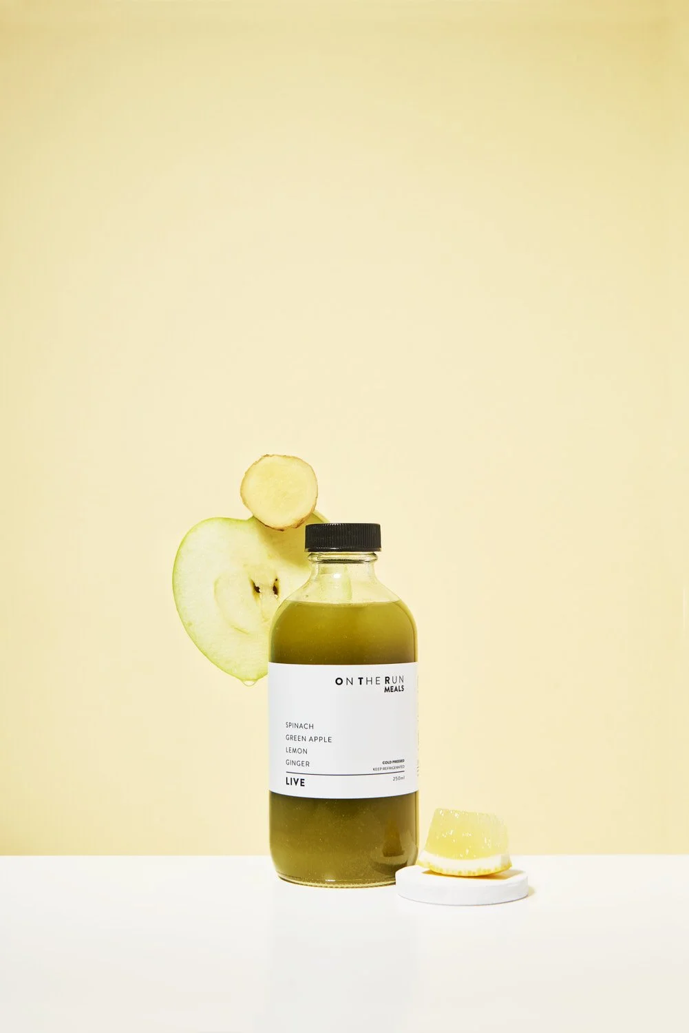 Single branded small juice or wellness shot bottle on a soft warm yellow background, clean minimal product photography with gentle side lighting