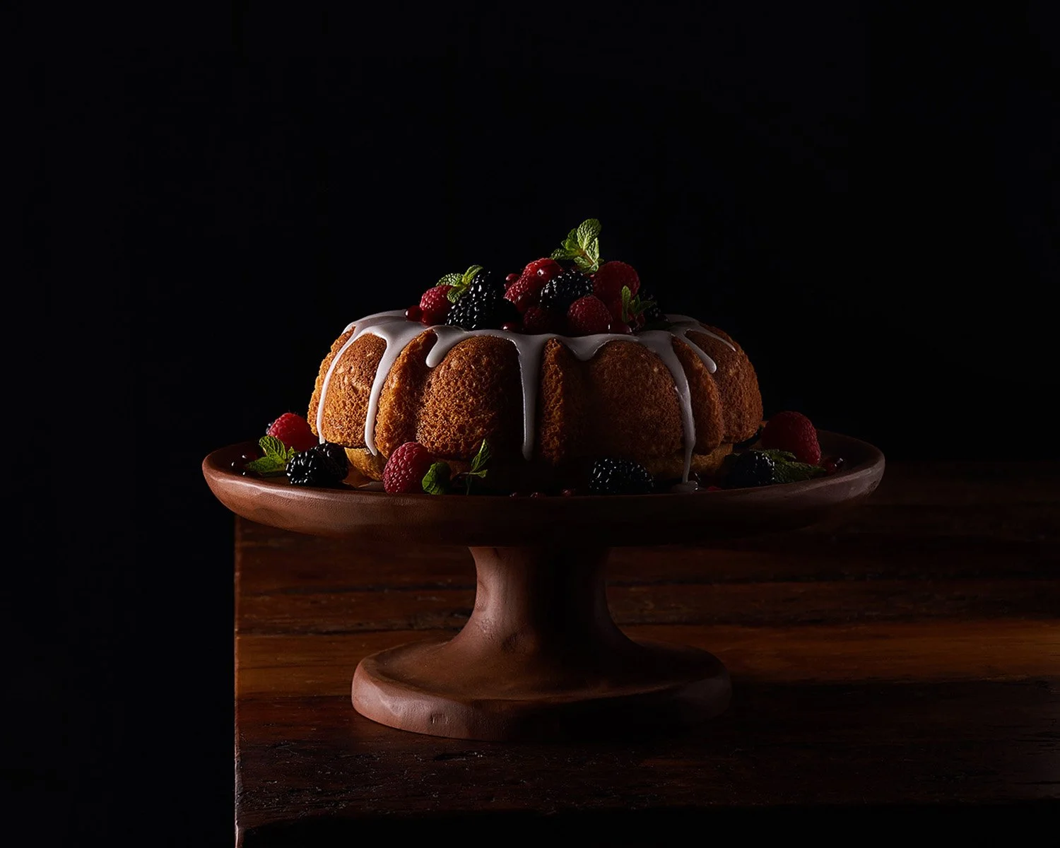 Moody low-key studio shot of a dark chocolate bundt cake on a raised pedestal, set against a deep black background with dramatic single-source lighting