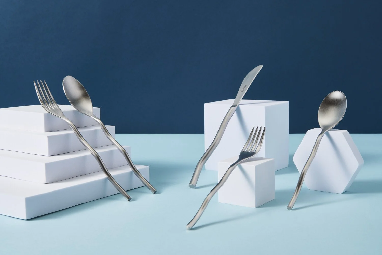 Artistic flat lay of white and silver cutlery and flatware arranged in a sculptural overlapping composition on a teal blue background with clean studio lighting