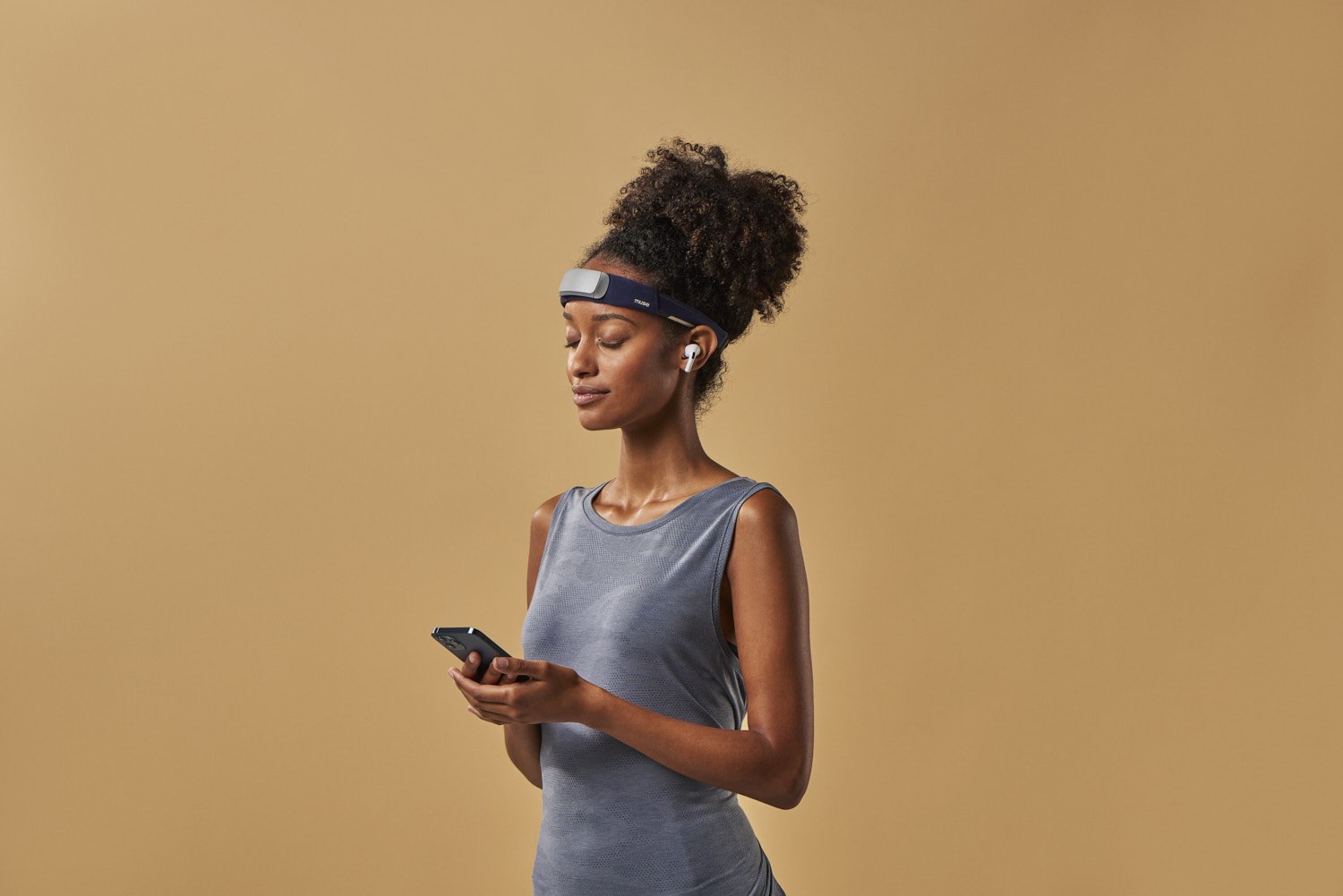 Young Black woman standing in profile looking at her phone while wearing smart glasses, photographed against a warm tan background with soft studio lighting