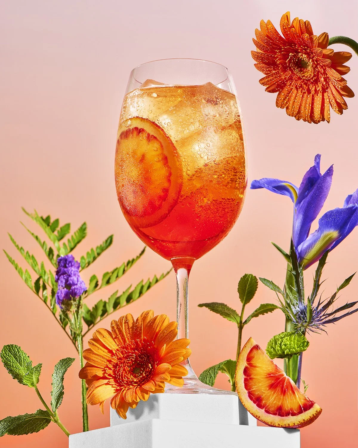 Glass of iced drink with orange slice, surrounded by colorful flowers and citrus wedge.