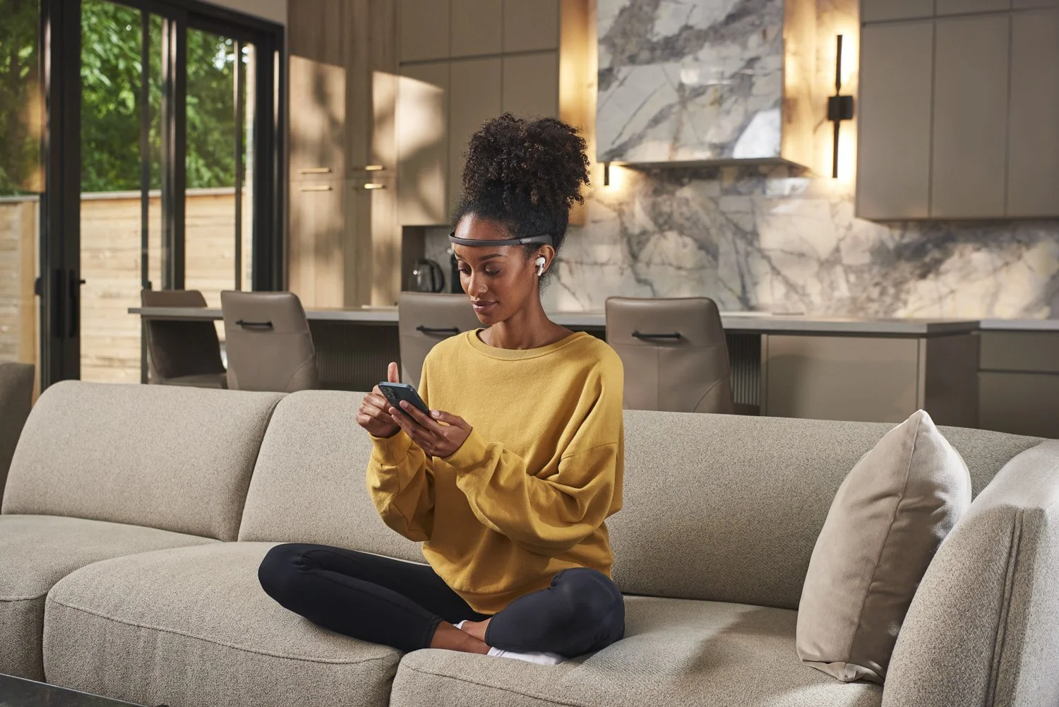 Young Black woman sitting cross-legged on a sofa looking at her phone, wearing a yellow top, warm lifestyle portrait with golden interior ambient lighting