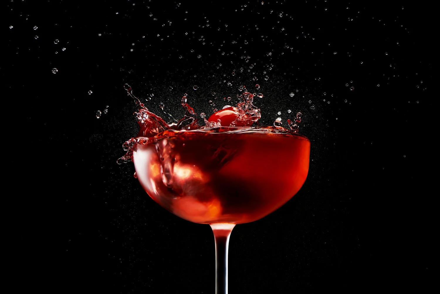 Dramatic dark studio shot of a deep red wine or cocktail in a coupe glass with liquid splashing dynamically against a pure black background