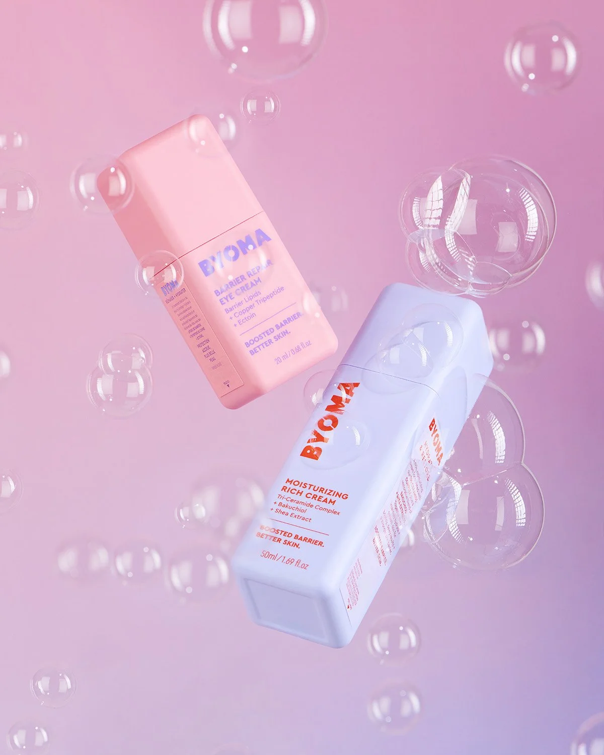 Two Byoma skincare bottles, one pink labeled Barrier Repair Eye Cream and one blue labeled Moisturizing Rich Cream, floating amidst bubbles on a pink background.