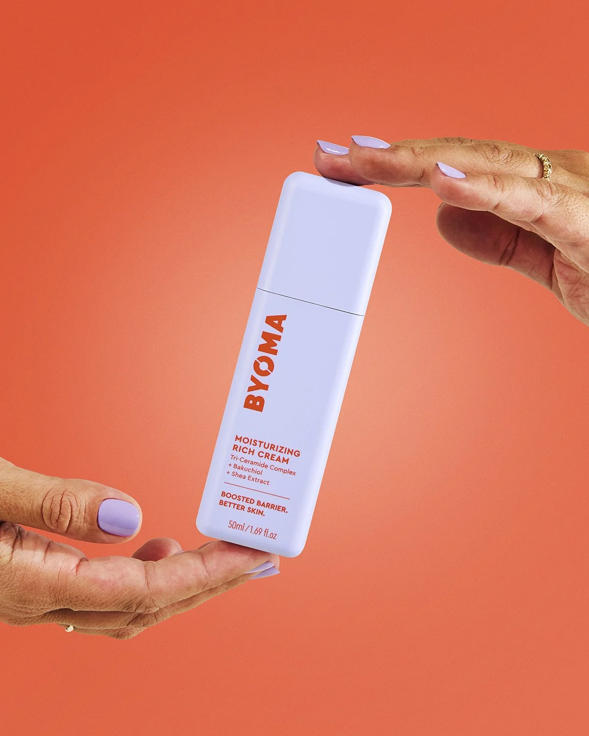 Hand holding a Byoma skincare tube vertically against a bold orange-red background, clean and confident product-in-hand commercial shot with strong color contrast