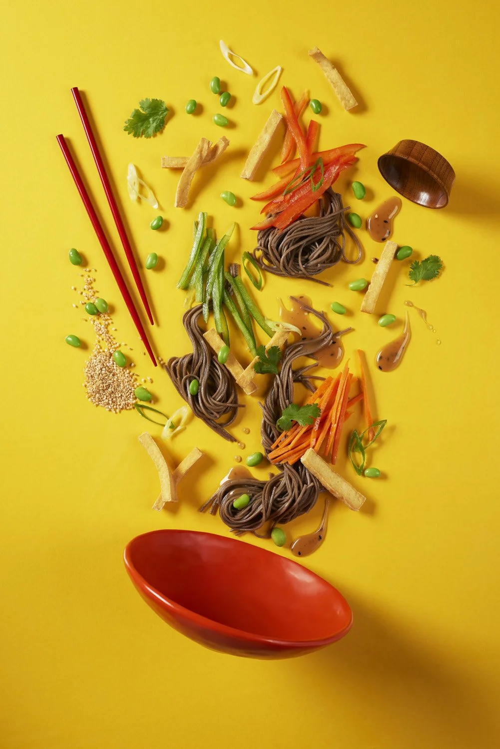 Arrangement of Asian-inspired ingredients on a yellow background, including noodles, sliced vegetables, green peas, tofu, sesame seeds, cilantro, chopsticks, and a red bowl.