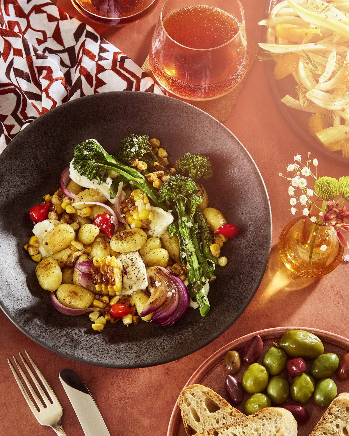 Grilled gnocchi and mixed olive salad in a wide bowl on a warm terracotta background, garnished with fresh herbs and a fork resting on the side