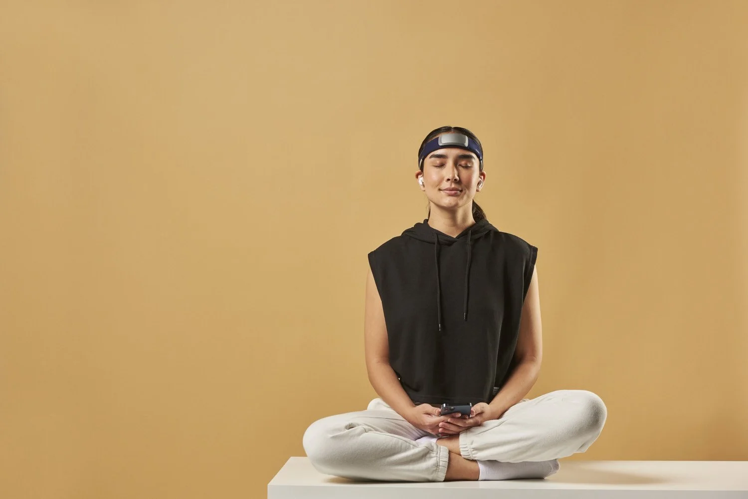 Young man in a black tank top sitting cross-legged in a meditative pose wearing a tech headband wearable, photographed against a warm golden tan background