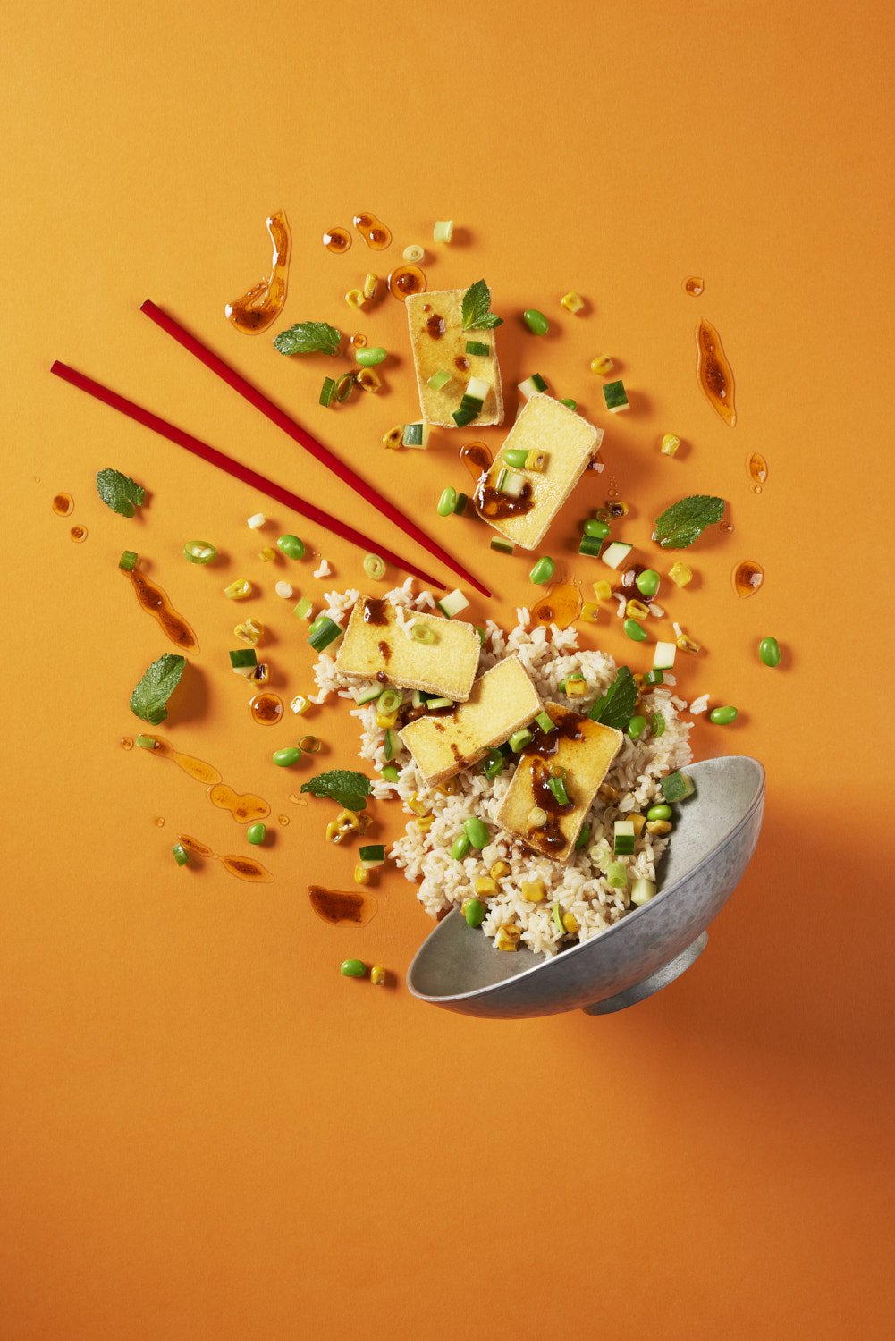Tossed rice bowl with colorful stir-fried vegetables and chopsticks resting across the rim, shot on a vivid orange background with strong overhead lighting