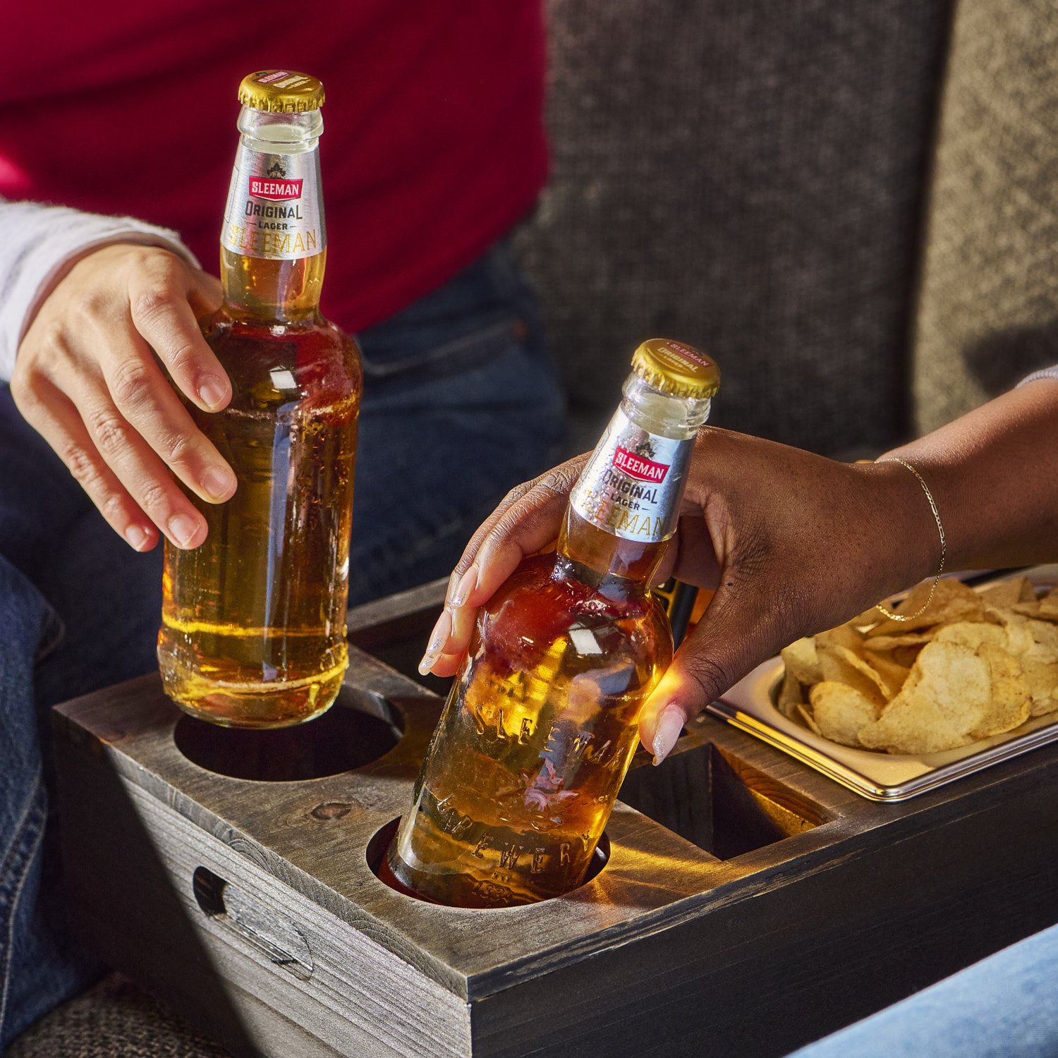 Person in a red sweater reaching into an open cooler to grab a glass bottle of beer, casual lifestyle shot with warm indoor lighting