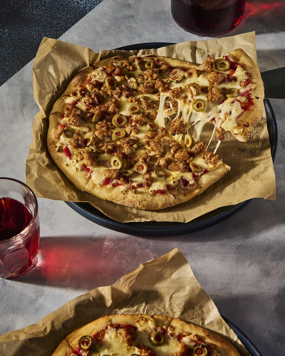 Rustic pizza loaded with toppings and melted cheese, photographed on a concrete surface alongside a glass of deep red wine