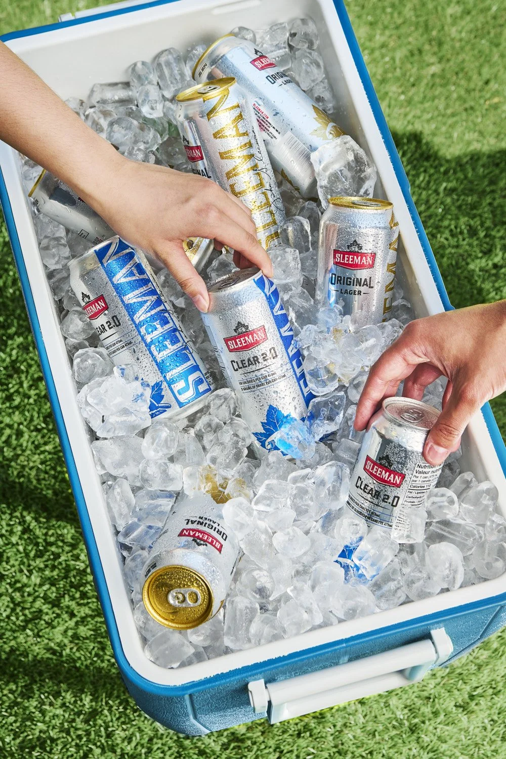 Hand reaching into an ice-filled cooler to grab a canned beverage, outdoor lifestyle shot on green grass with natural daylight from above