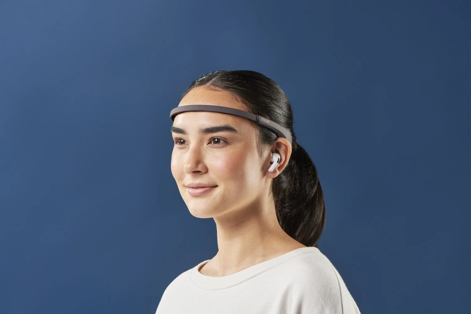 Side profile of a young woman wearing a slim tech headband wearable device, shot against a solid dark blue background with clean studio lighting