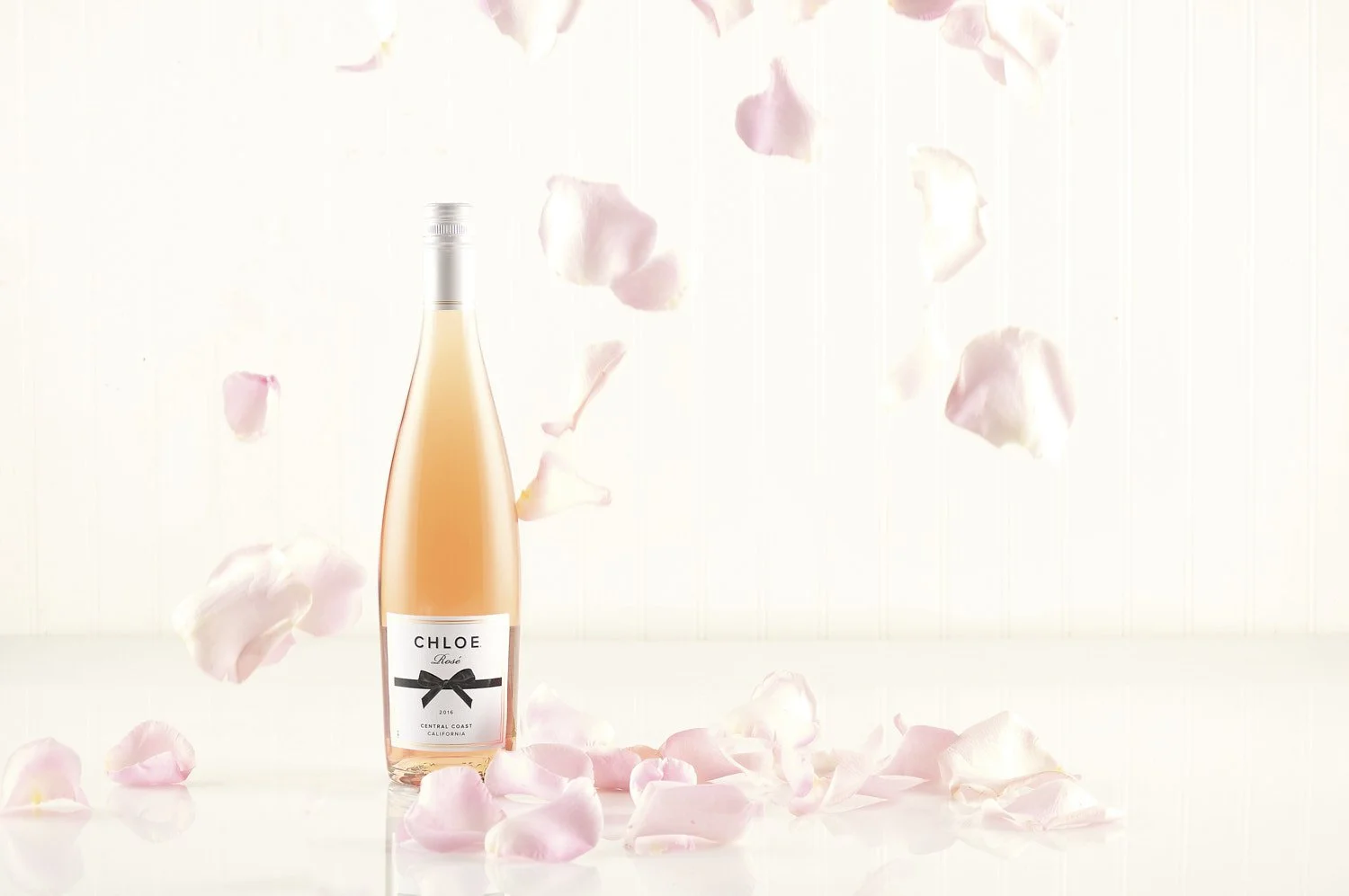 Delicate rosé wine bottle with petals and small flowers floating and falling around it, ethereal light and airy product shot on a soft white background