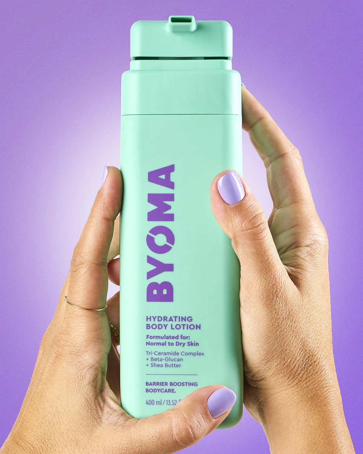 Hand holding a tall green Byoma skincare bottle upright against a vibrant purple background, bold product-in-hand commercial shot with strong vertical composition