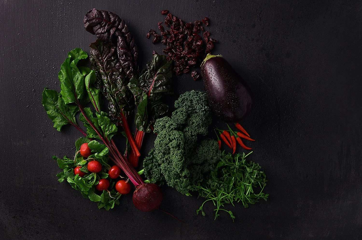 Dark moody arrangement of roasted broccolini, purple kale, and colorful vegetables spread across a black slate surface with dramatic low-key studio lighting