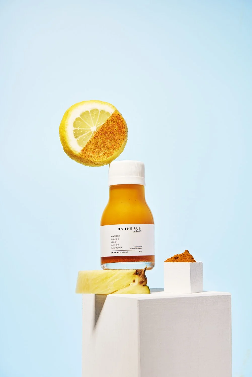 Branded juice bottle with a halved orange slice suspended mid-air beside it, shot on a crisp sky blue background with a white pedestal base