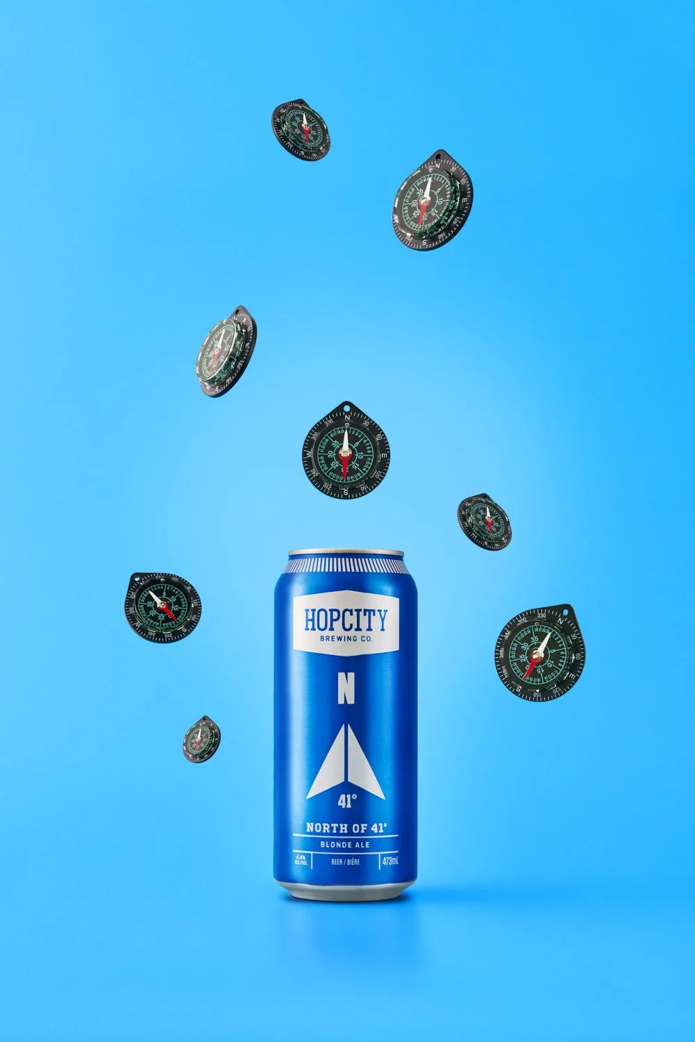 Branded beer can on a bold blue background with dark coffee beans and seeds scattered and falling dynamically around the product