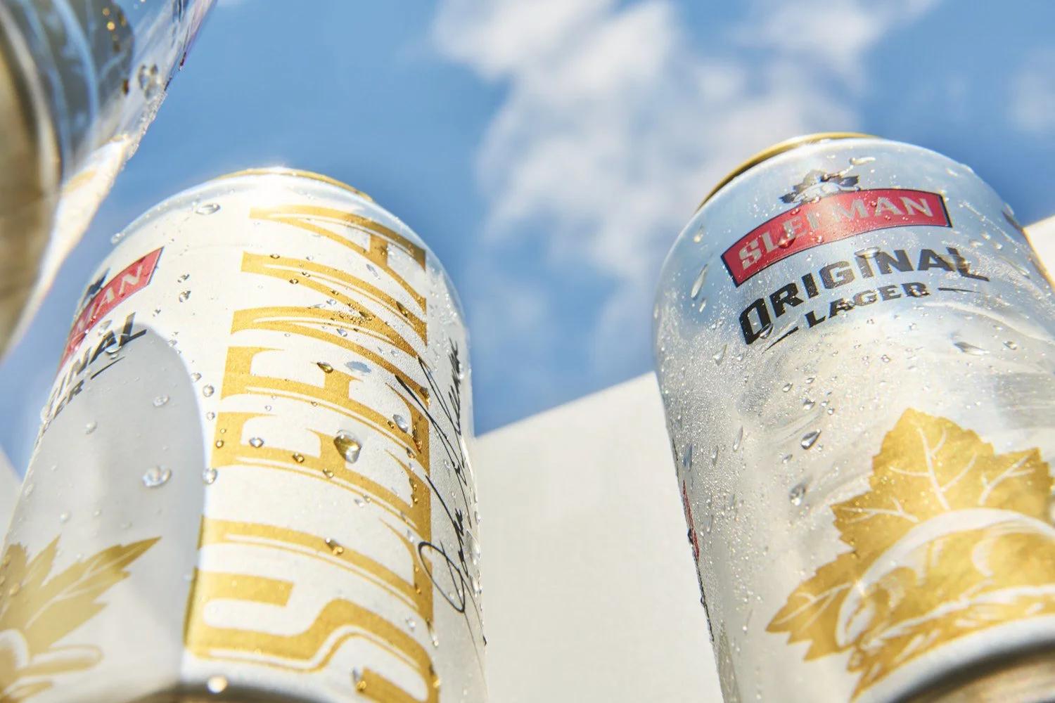 Close-up of a branded aluminum beverage can held against a bright blue sky with soft clouds, strong natural light and upward angle