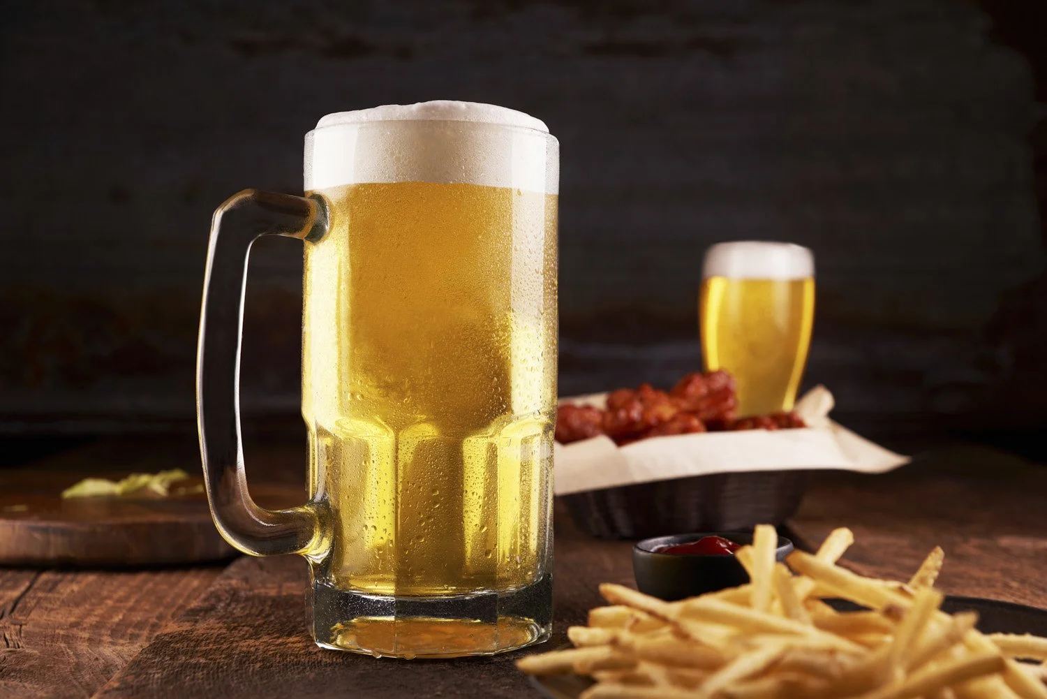  tall frosted glass of golden beer beside a plate of crispy salted french fries, shot on a rustic wooden surface with warm ambient lighting