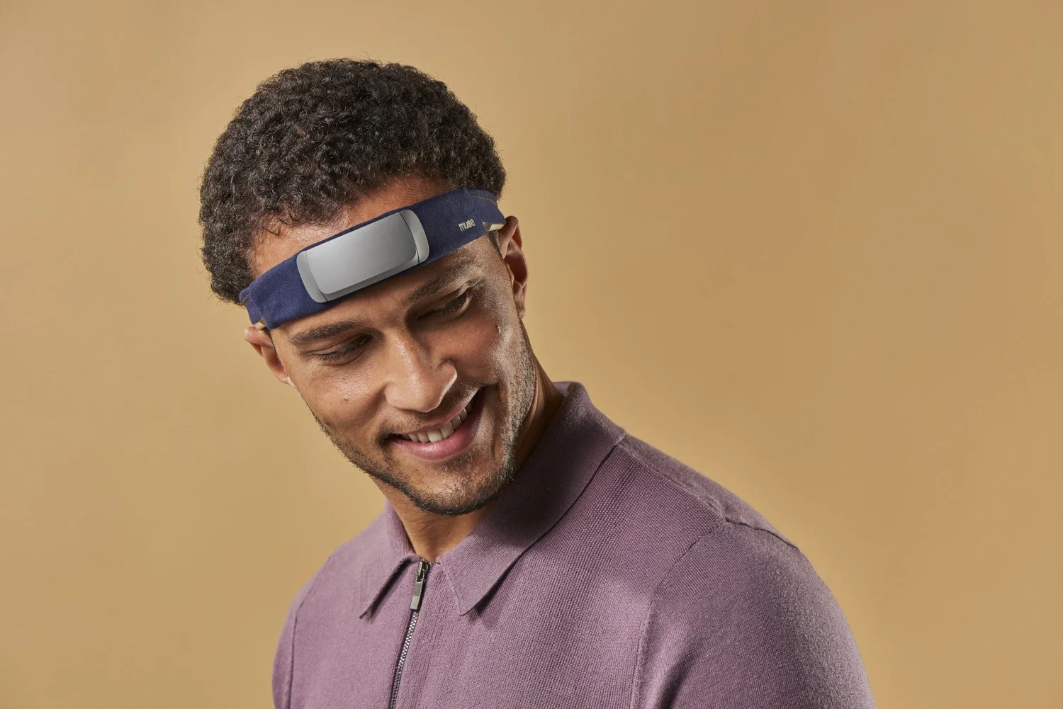Young man smiling while wearing a tech headband wearable device, shot against a warm beige background with soft directional studio lighting and relaxed expression