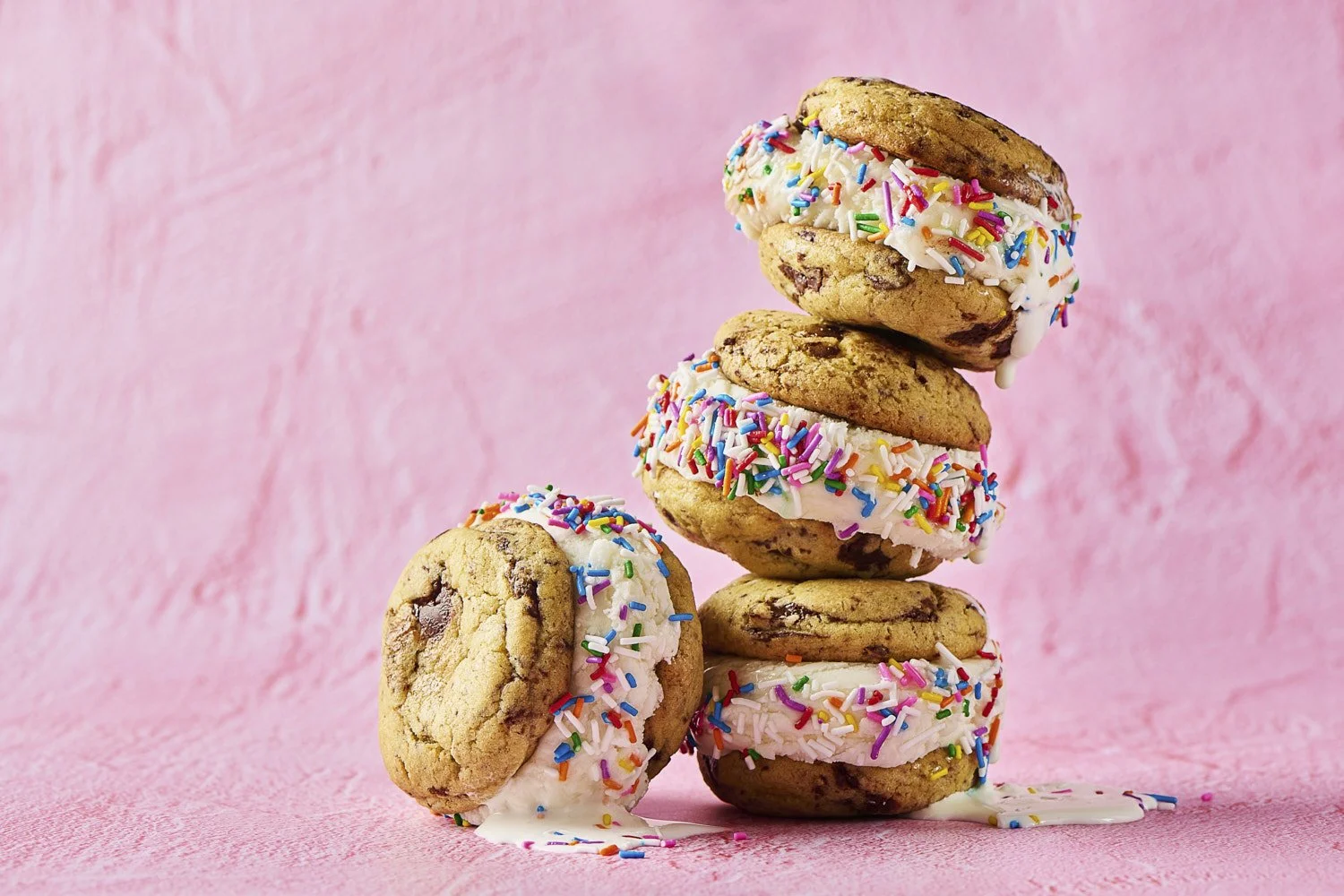 Stack of three colourful sprinkle-frosted ice cream sandwiches leaning against each other on a bright pink background, playful pastel food styling with soft even lighting