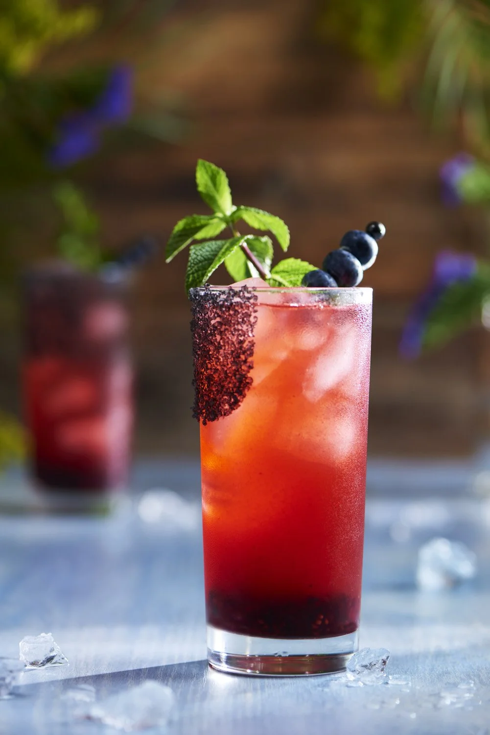 all red cocktail garnished with fresh mint and berries in a highball glass, shot on a soft blue surface with scattered botanicals and warm side lighting