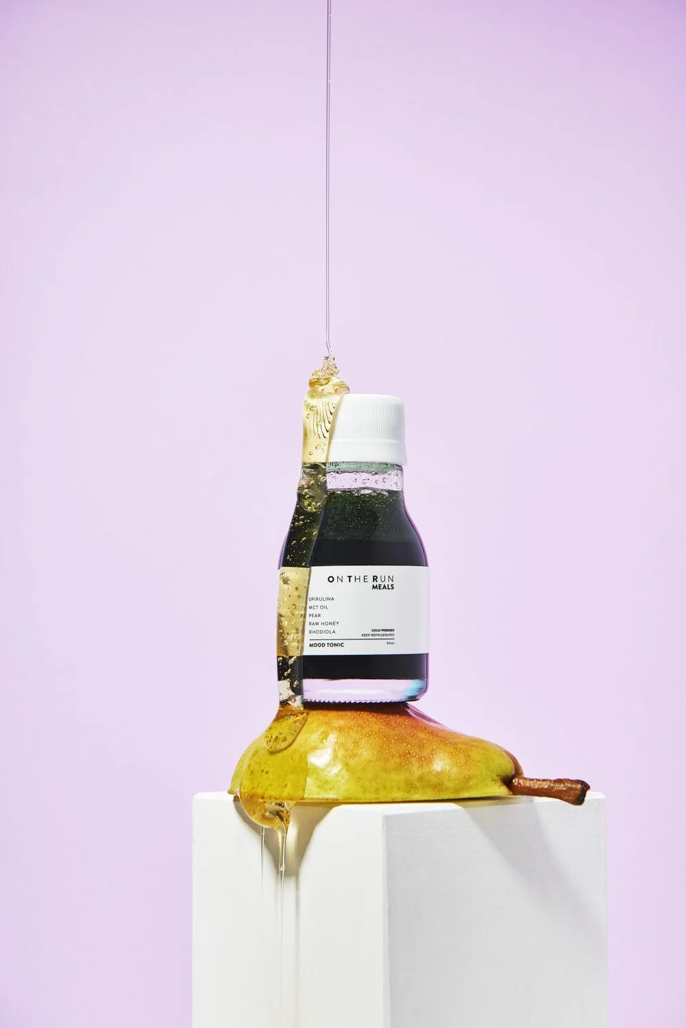 Dark glass syrup or liqueur bottle standing on a ripe pear against a soft pastel pink and purple split background, minimal product styling with even light