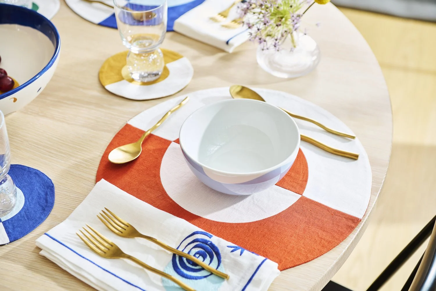 Elegantly styled dining table with white and blue patterned plates, gold cutlery, red placemats, and floral centerpiece, shot from above in warm natural light