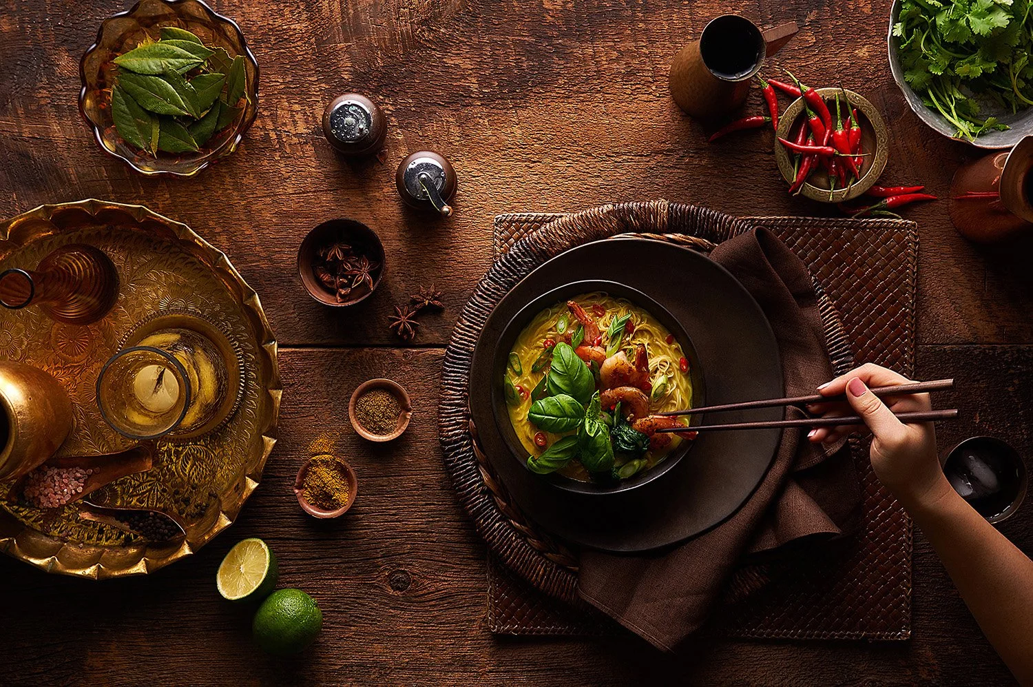 Overhead rustic shot of a dark stew or curry in a wide earthenware bowl on a wooden table, surrounded by small condiment dishes and garnishes