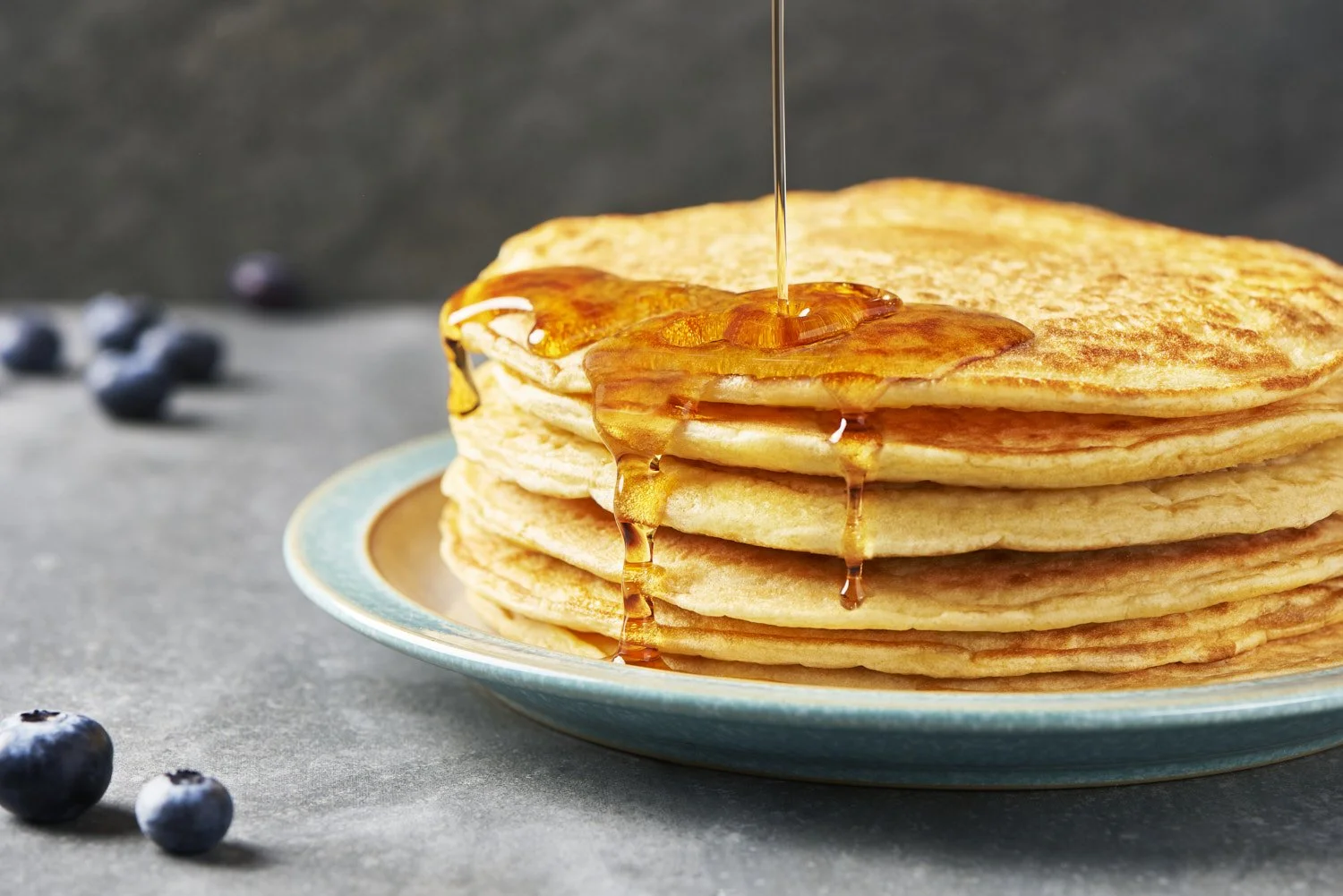 Tall stack of thin golden French-style pancakes with honey being drizzled from above, fresh blueberries scattered alongside on a clean white serving plate
