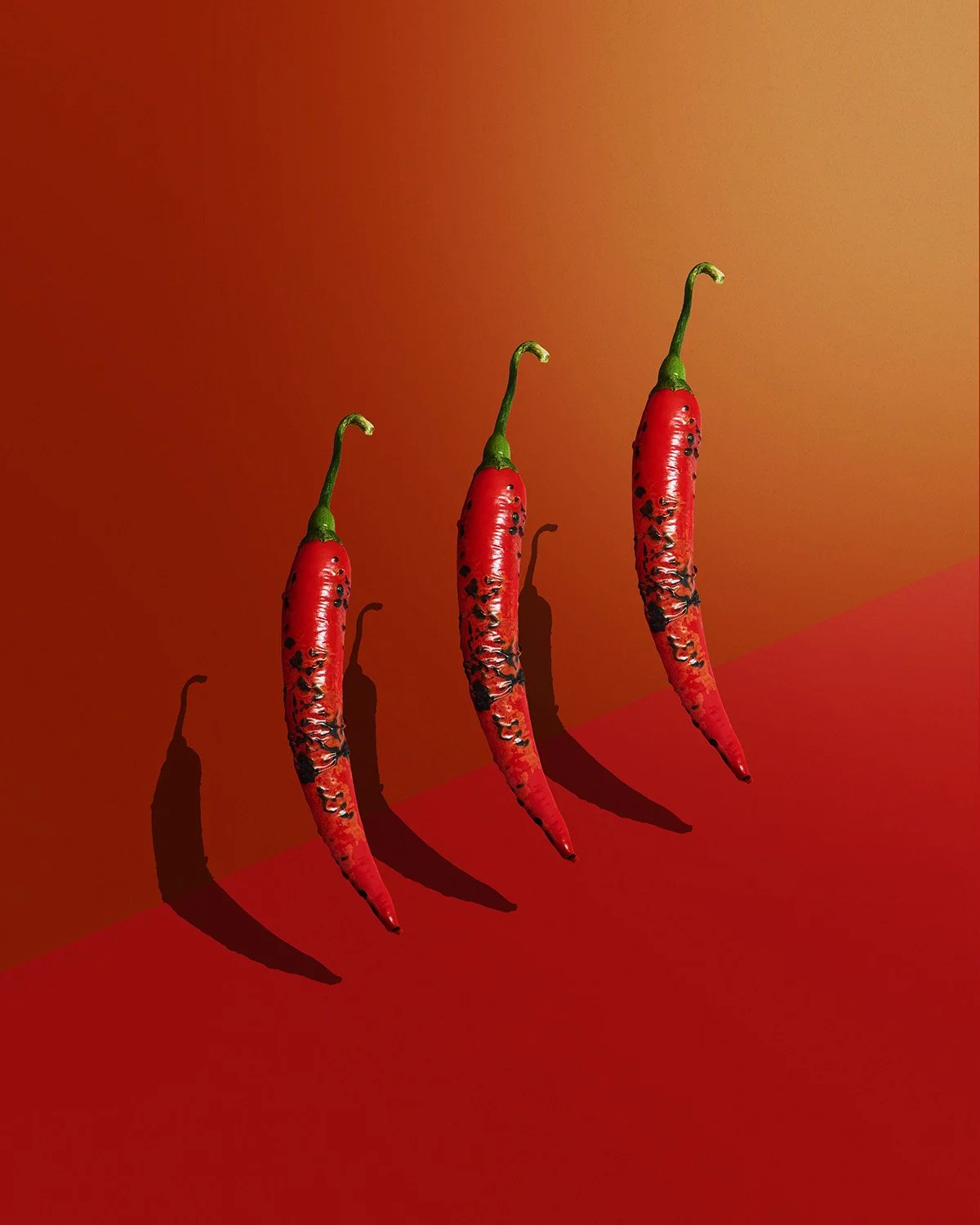  Three long red cayenne chili peppers arranged diagonally against a deep saturated red background, strong directional side lighting creating bold shadow and texture contrast