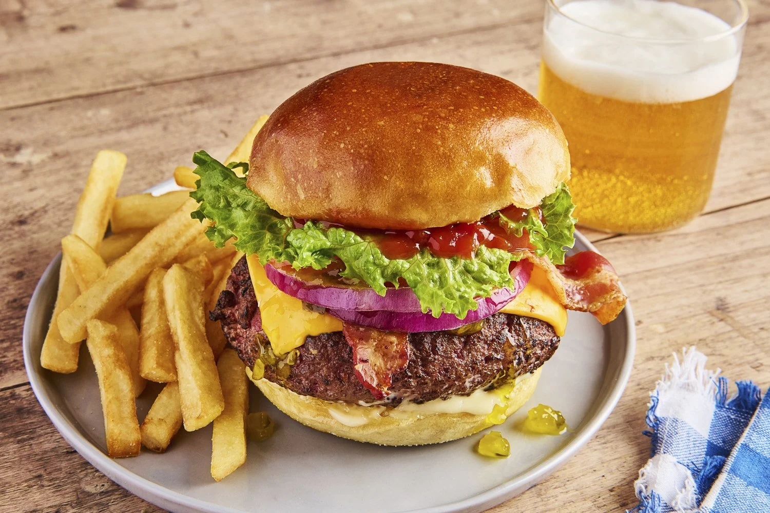 Tall stacked gourmet burger with lettuce, pickles, and condiments on a brioche bun, served with golden fries and a cold beer on a bright clean surface