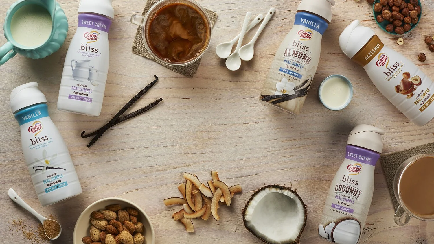 Overhead flat lay of branded coffee creamer bottles surrounded by vanilla pods, coconut, nuts, and spices on a warm light wooden surface