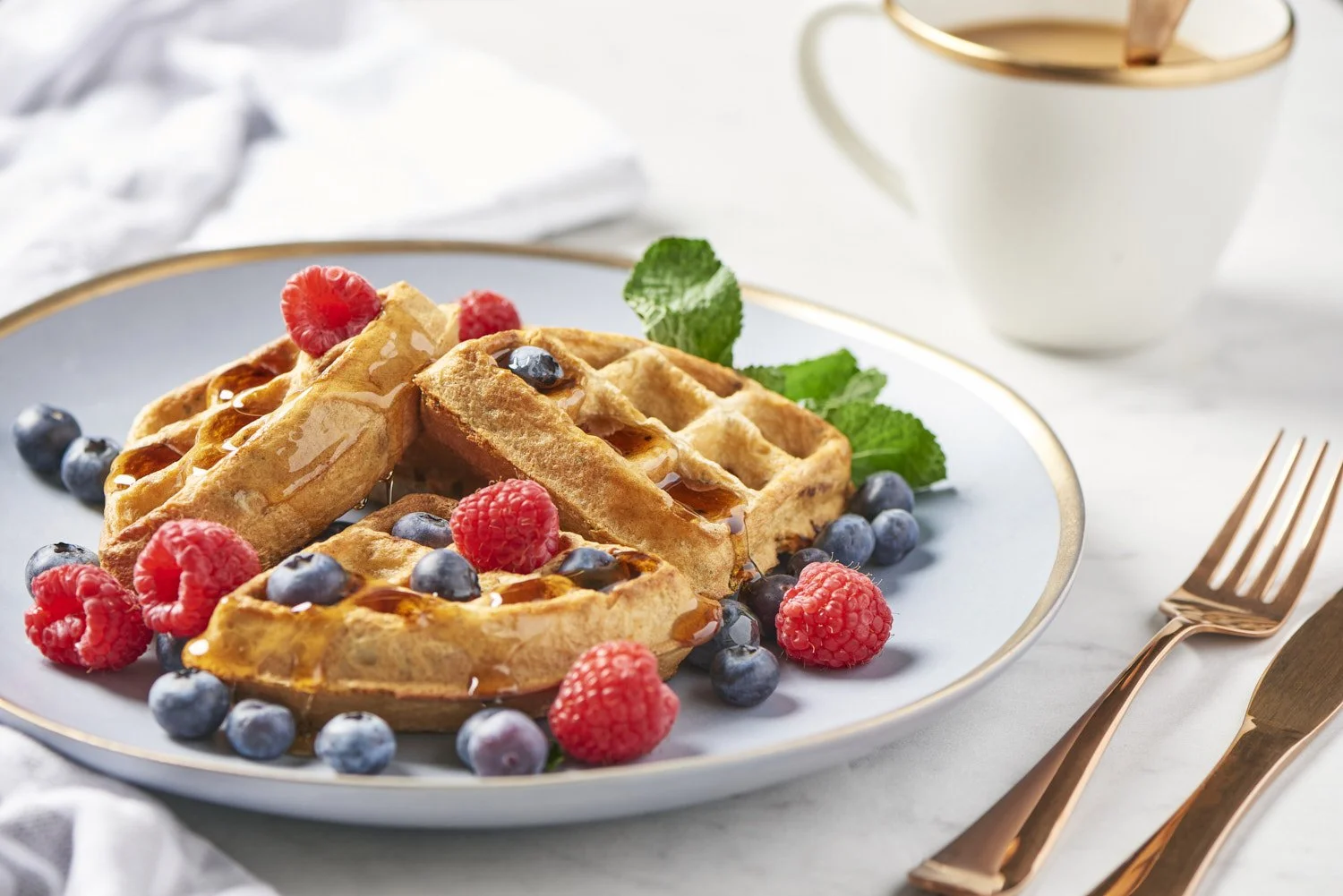 Belgian waffles topped with fresh blueberries, raspberries, and a dollop of whipped cream, served on a white plate with a coffee mug in the background