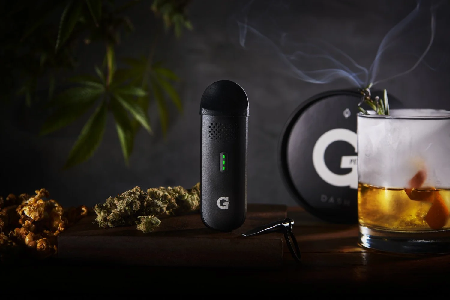 Dark moody product shot of a black vape  beside a whiskey glass, low-key studio lighting with tropical leaves and smoke in the background