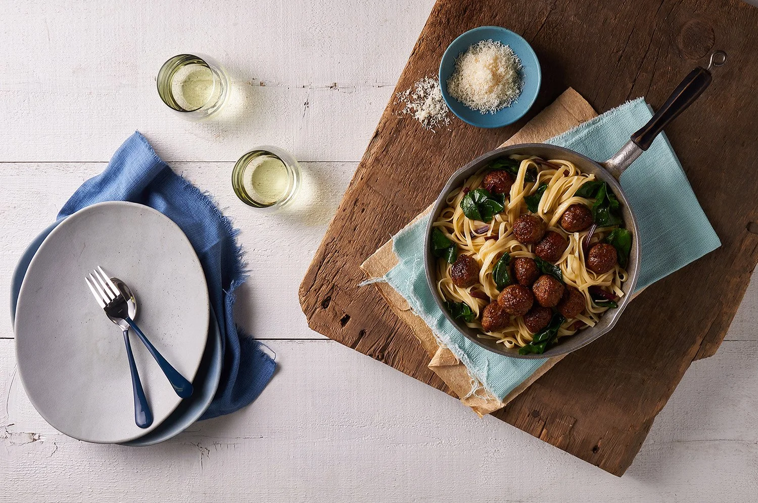 Overhead shot of pasta with braised dark leafy greens and meatballs served in a wide white bowl, with a light linen napkin and small sauce dish alongside