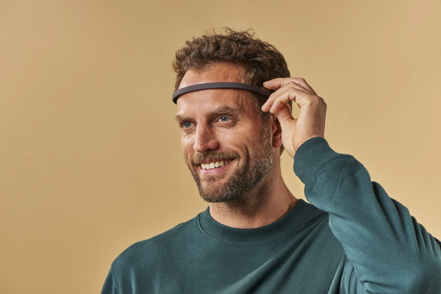 Man in a green shirt adjusting a slim tech headband wearable on his temple, shot against a warm tan background with natural lifestyle expression