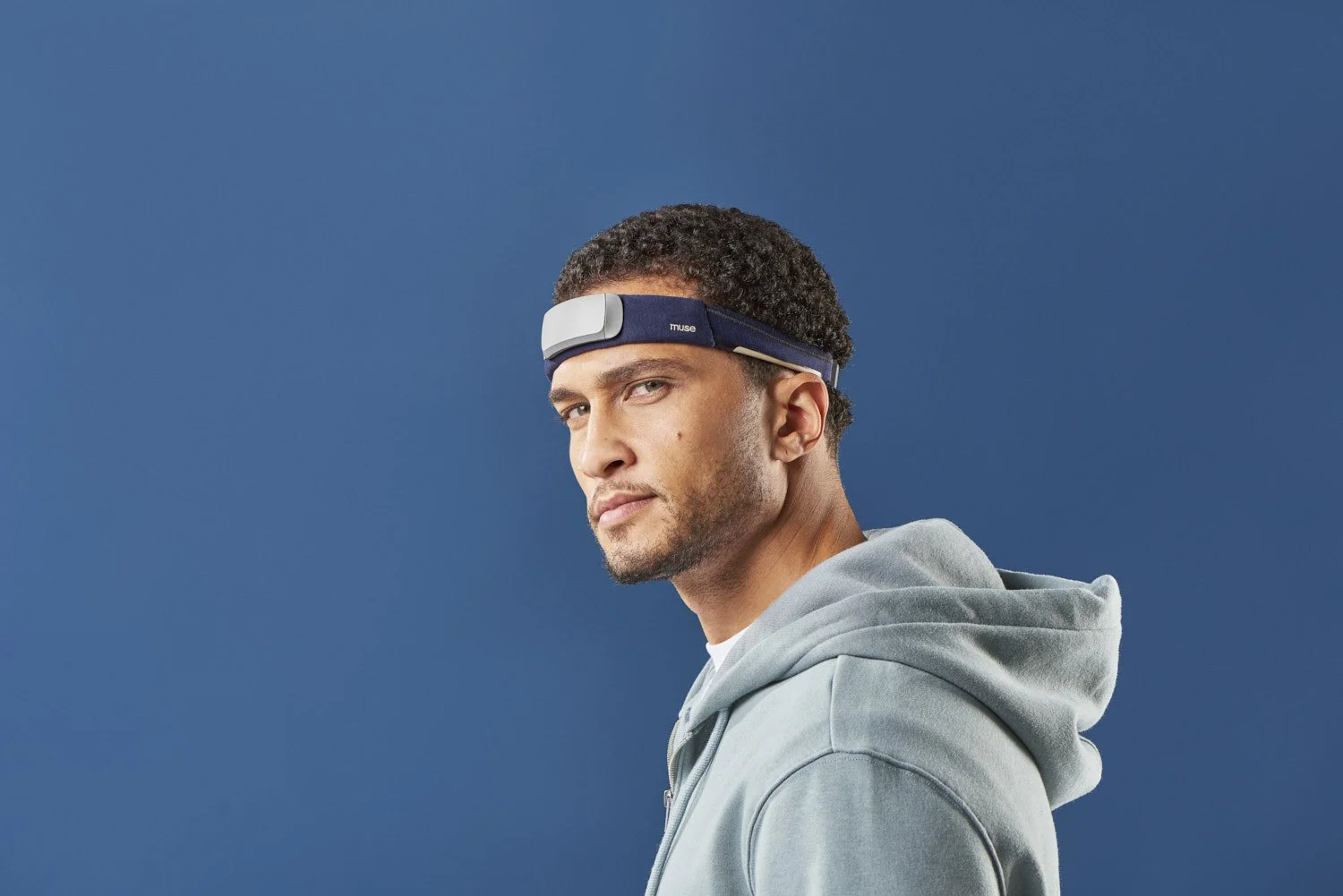 Side profile of a young man in a grey hoodie wearing a tech headband wearable device, shot against a solid dark blue background with clean lighting
