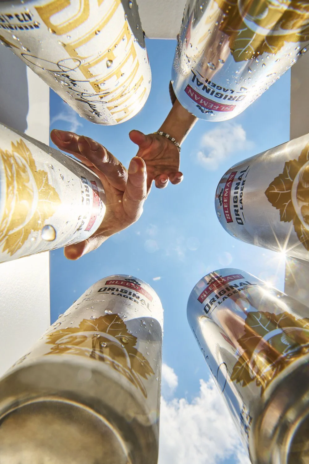 Worm's-eye view looking up at multiple cocktail glasses held together in a toast, shot from below against a bright blue sky for dramatic perspective