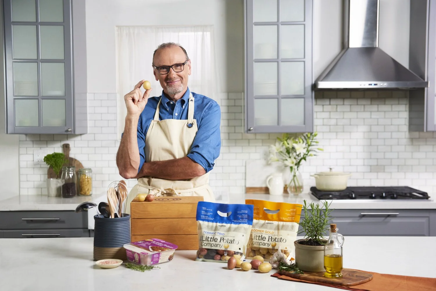 Chef Michael Bonacini in an apron standing at a kitchen counter with branded food products, herbs, and cooking ingredients, warm lifestyle commercial photography with natural light