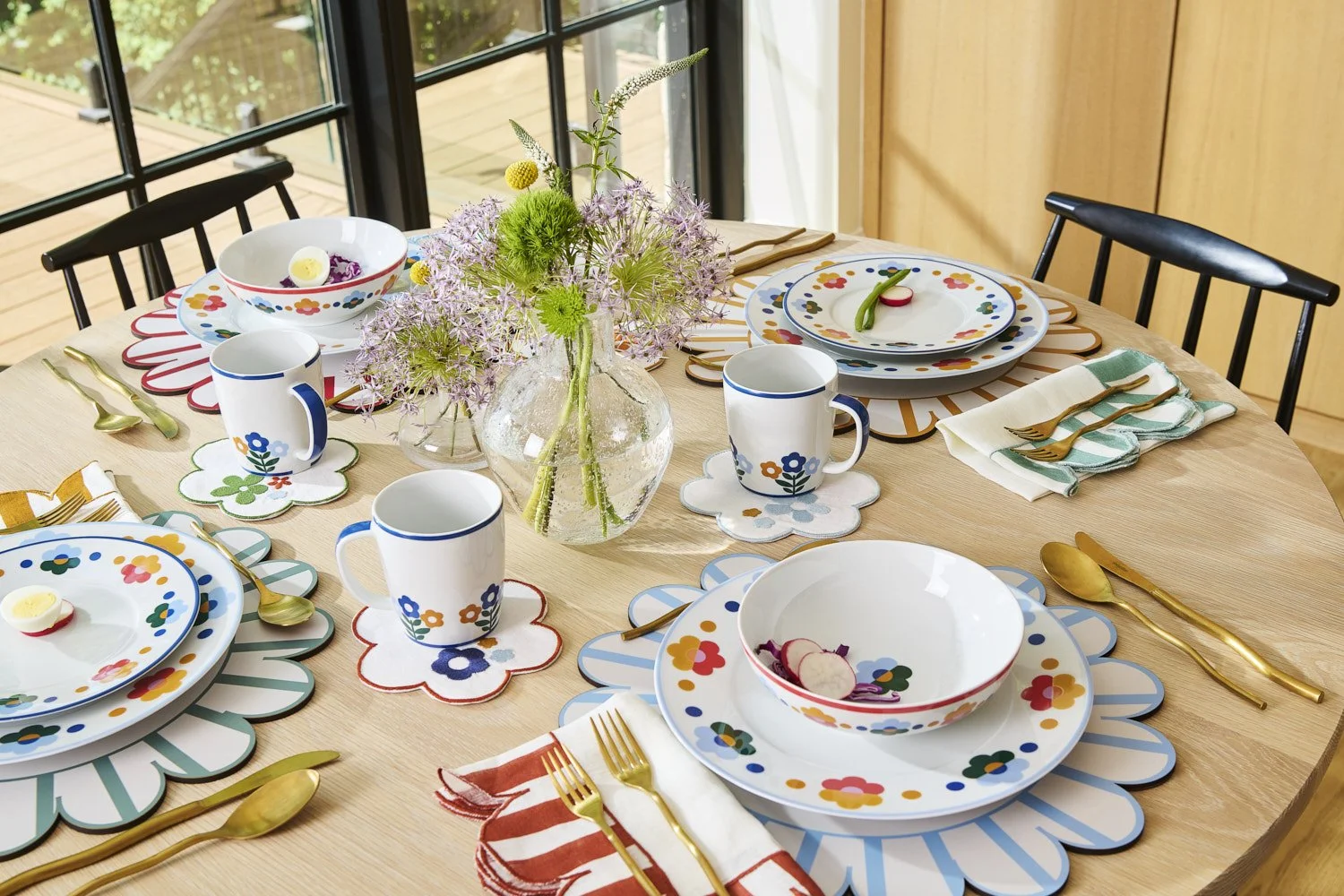 Overhead view of a beautifully set dining table with colorful patterned plates, glassware, and floral centerpiece, warm natural light from a large window nearby
