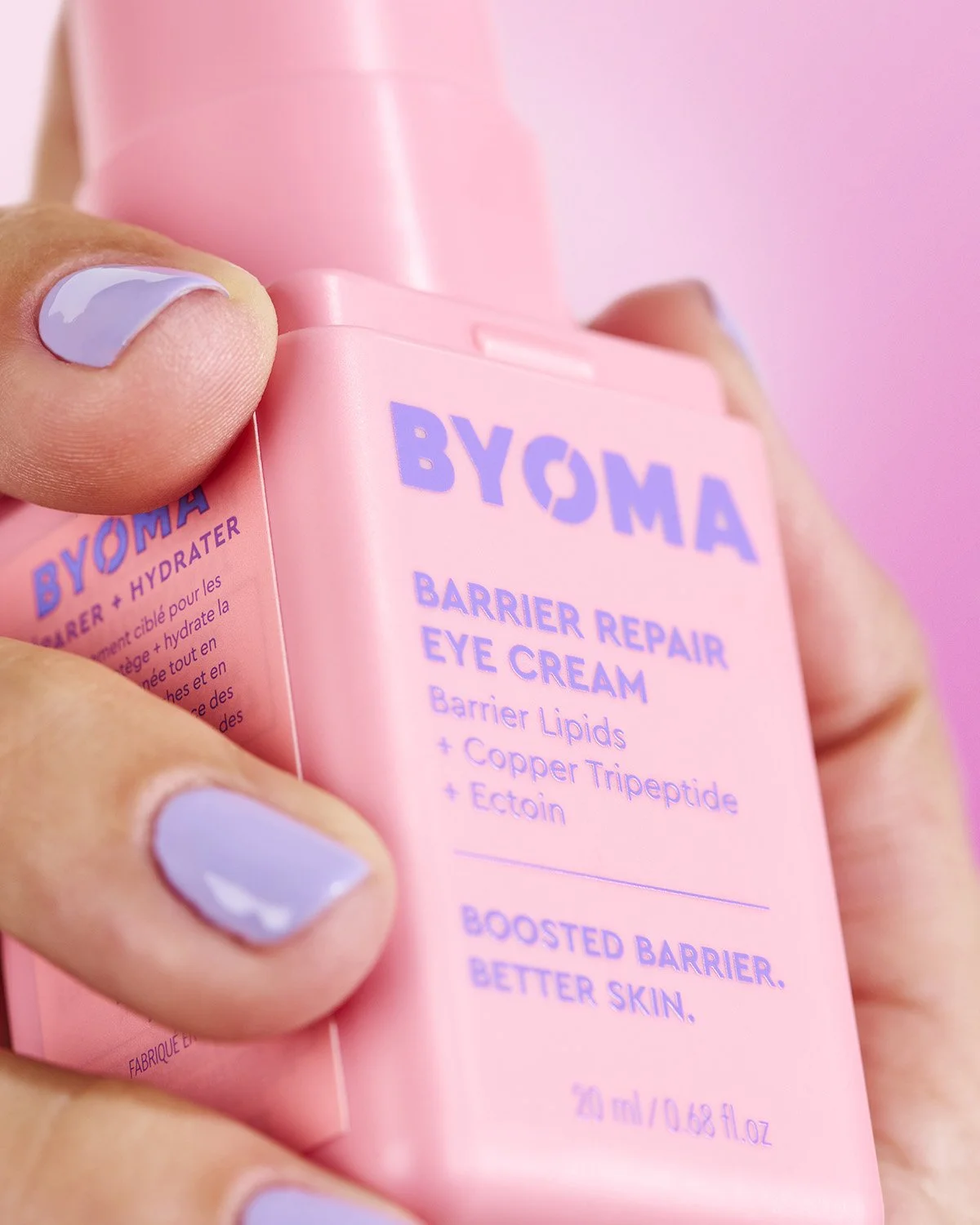 Close-up of a hand with lavender nail polish holding a Byoma Barrier Repair Eye Cream tube against a soft pink background, product label clearly visible