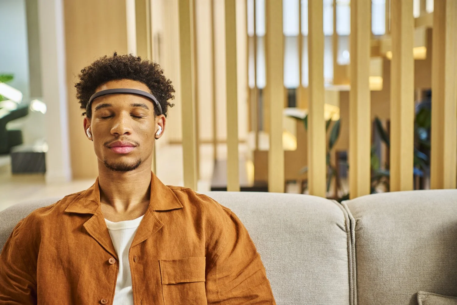 Young Black man seated on a modern sofa in a warmly lit contemporary interior, wearing a sleek headband tech wearable, lifestyle portrait with natural expression