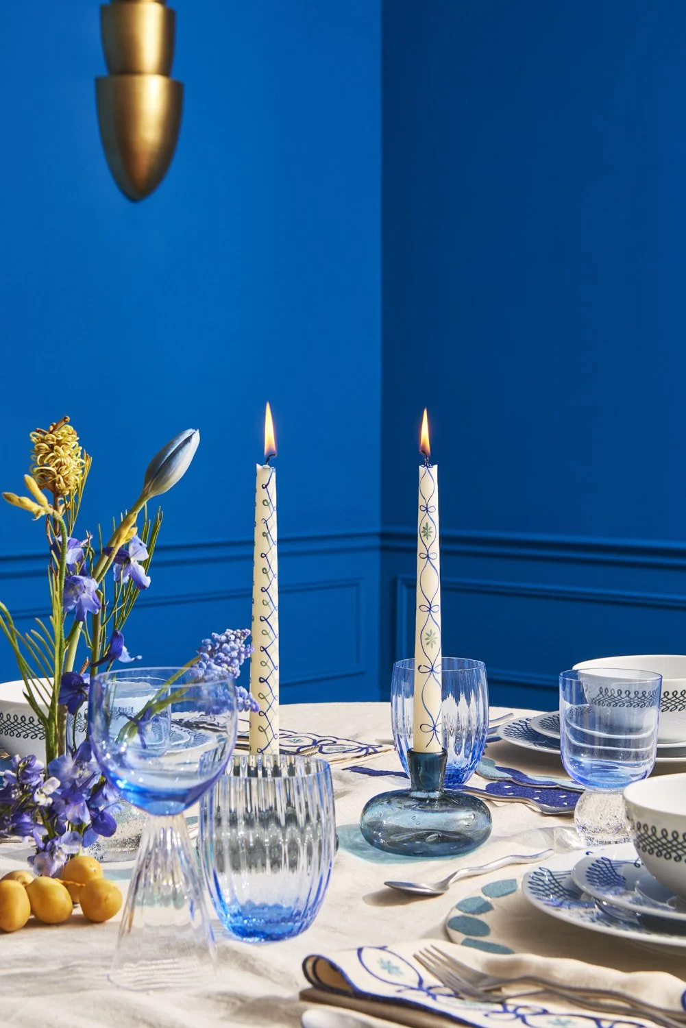 Elegantly set dining table with blue glassware, tall taper candles, blue plates, and floral arrangements, moody lifestyle tablescape shot against a deep blue wall